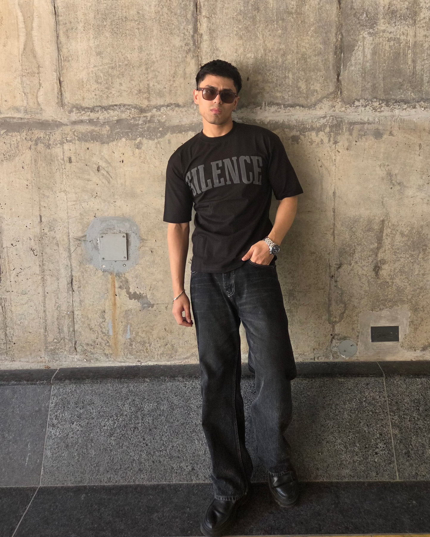 Person wearing a black t-shirt with 'Silence' printed on it, standing against a stone wall.