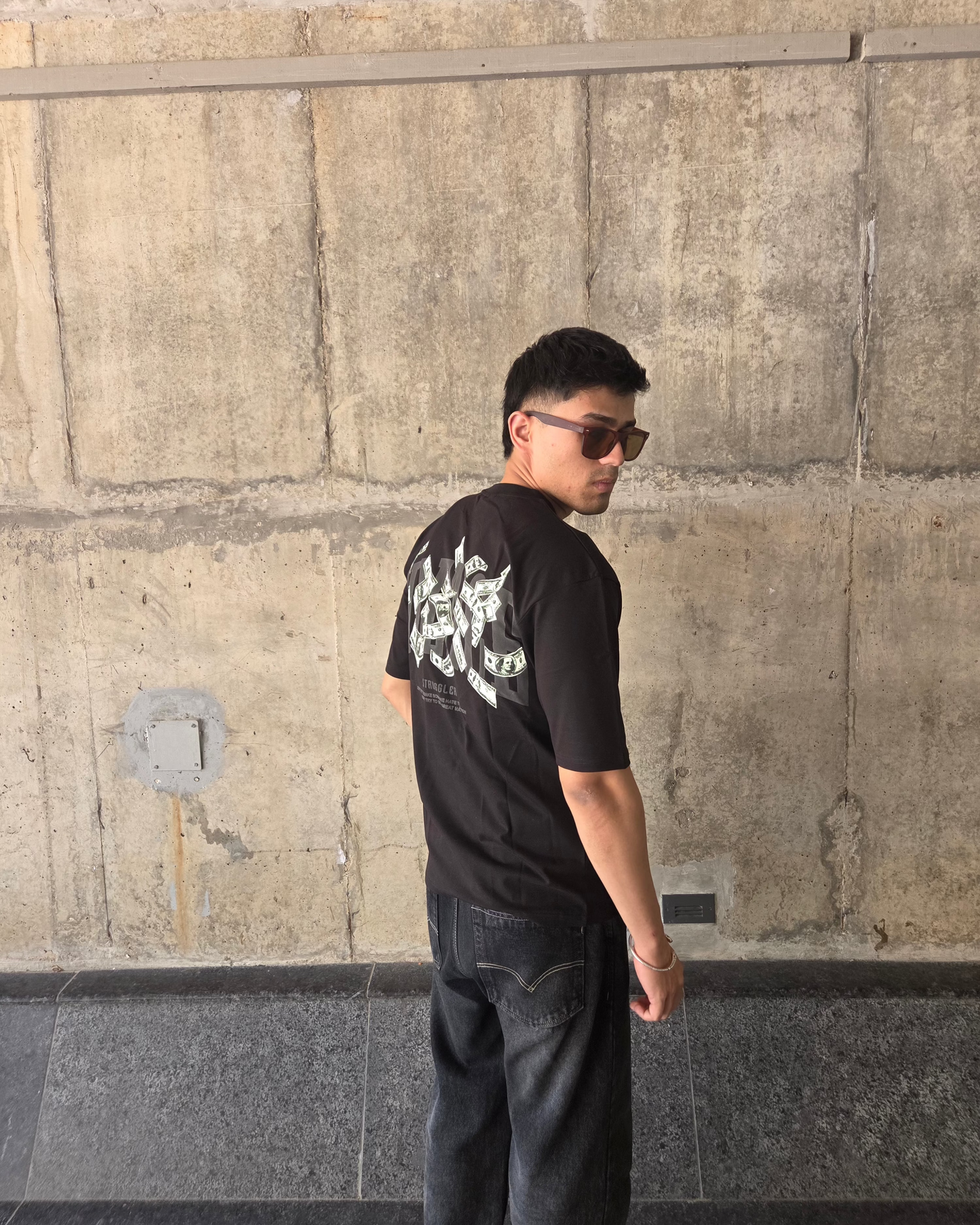 Man wearing a black t-shirt with a graphic design and sunglasses, standing against a concrete wall.